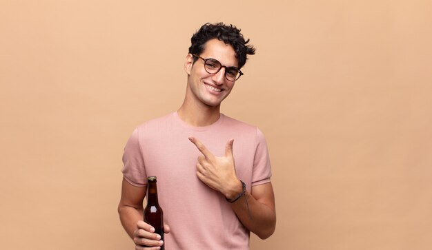 Hombre joven con una cerveza sonriendo alegremente, sintiéndose feliz y apuntando hacia un lado y hacia arriba, mostrando el objeto en el espacio de la copia