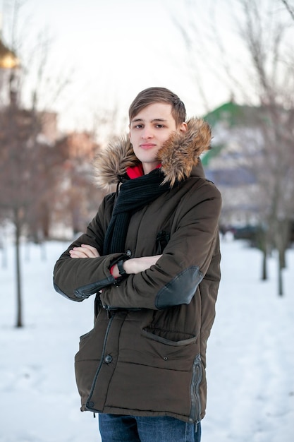 Un hombre joven con una chaqueta de invierno con pieles se encuentra con los brazos cruzados en un parque