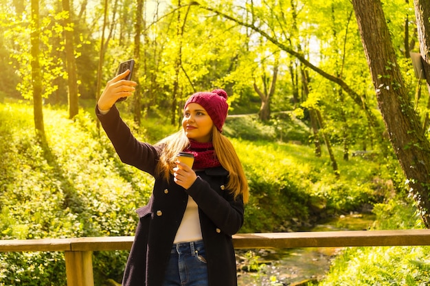 Foto hombre joven con una chaqueta negra, bufanda y gorro de lana rojo disfrutando en un parque de otoño