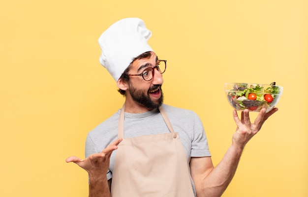 Hombre joven chef barbudo sorprendido expresión y sosteniendo una ensalada