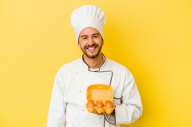 Hombre joven chef caucásico con huevos aislados sobre fondo amarillo feliz, sonriente y alegre.