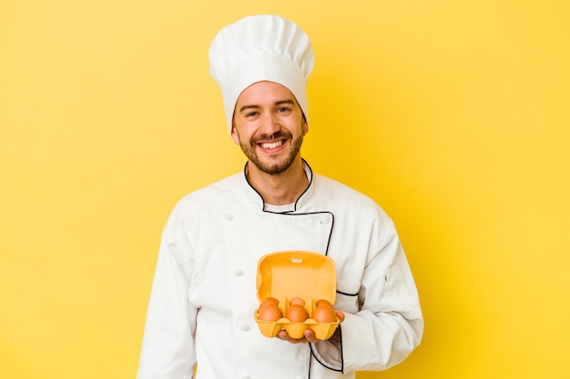 Hombre joven chef caucásico con huevos aislados sobre fondo amarillo feliz, sonriente y alegre.