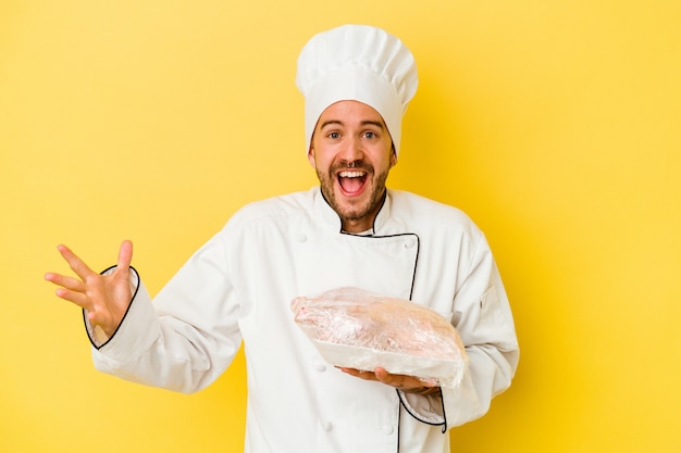 Hombre joven chef caucásico sosteniendo pollo aislado sobre fondo amarillo recibiendo una agradable sorpresa, emocionado y levantando las manos.