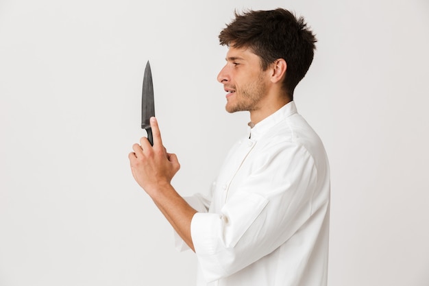 Hombre joven chef de pie aislado en la pared blanca con cuchillo.