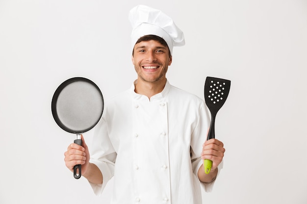 Hombre joven chef de pie aislado en la pared blanca sosteniendo una sartén.