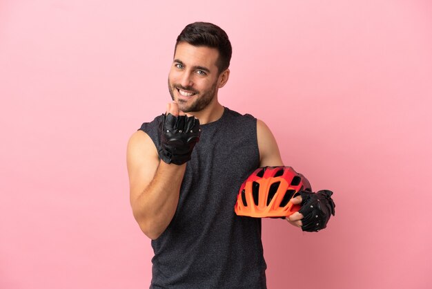 Hombre joven ciclista aislado sobre fondo rosa haciendo gesto de dinero