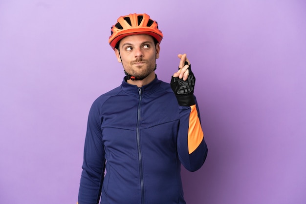 Hombre joven ciclista brasileño aislado sobre fondo púrpura con los dedos cruzando y deseando lo mejor