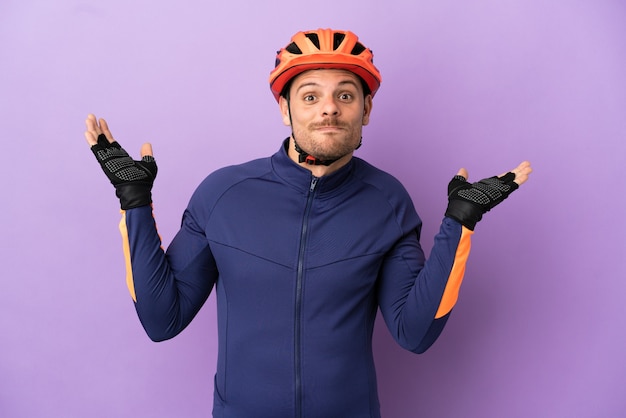 Hombre joven ciclista brasileño aislado sobre fondo púrpura con dudas mientras levanta las manos