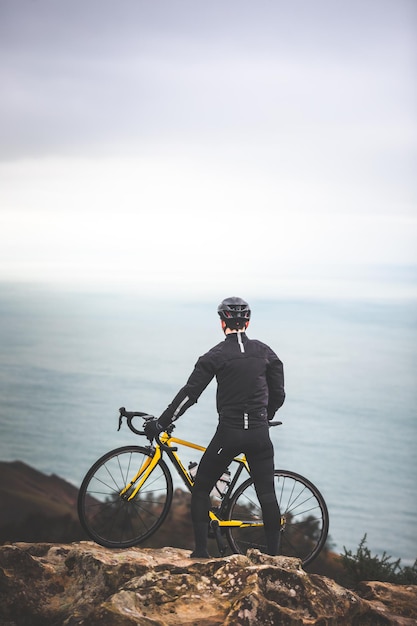 Hombre joven ciclista caucásico en la cima de la montaña mirando al mar.