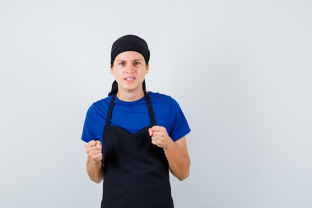 Foto hombre joven cocinero levantando los puños cerrados en camiseta, delantal y mirando furioso, vista frontal.