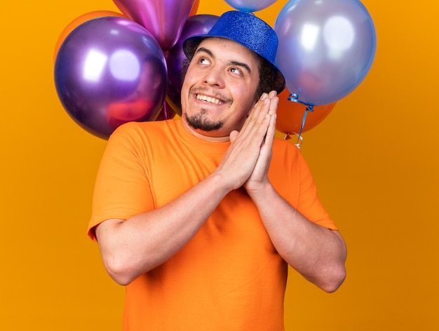 Hombre joven codicioso con sombrero de fiesta de pie delante de globos tomados de la mano juntos aislado en la pared naranja