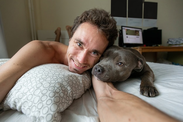 Hombre joven complacido mirando a cámara y tomando selfie con perro amstaff mientras está acostado en la cama