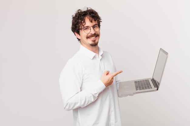 Hombre joven con una computadora portátil sonriendo alegremente, sintiéndose feliz y apuntando hacia un lado