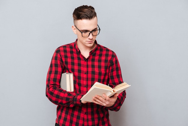 Hombre joven concentrado en gafas leyendo un libro en el estudio. Fondo gris aislado