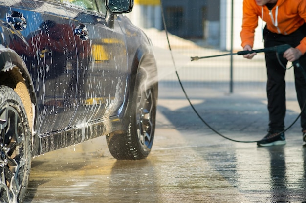 Hombre joven conductor lavando su coche con chorro de agua a alta presión sin contacto en autoservicio de lavado de coches.