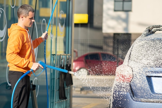 Hombre joven conductor lavando su coche con chorro de agua a alta presión sin contacto en autoservicio de lavado.