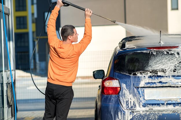 Hombre joven conductor lavando su coche con chorro de agua a alta presión sin contacto en autoservicio de lavado.