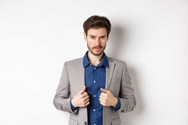 Hombre joven confiado con traje de arreglo de barba y mirando descarado a la cámara, sintiéndose listo y decidido, de pie sobre fondo blanco.