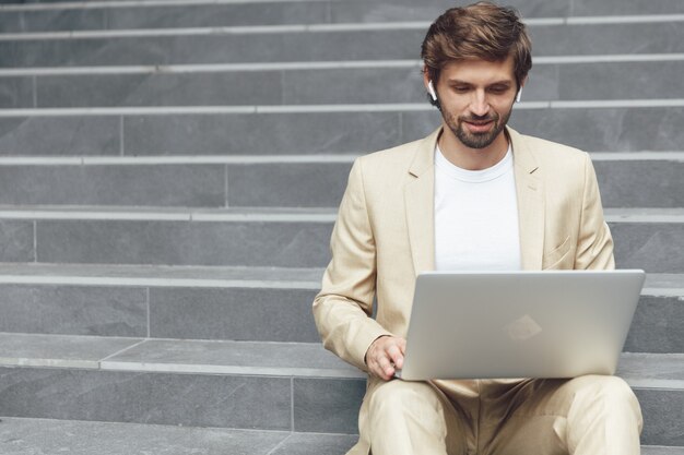 Hombre joven confidente en traje de negocios usando laptop portátil para trabajar mientras está sentado en las escaleras al aire libre