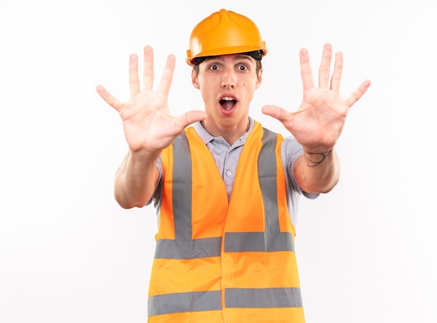 Hombre joven constructor asustado en uniforme mostrando gesto de parada aislado en la pared blanca