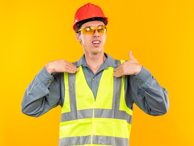 Hombre joven constructor disgustado en uniforme con gafas apunta a sí mismo