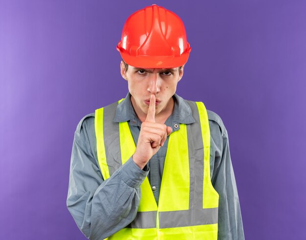 Hombre joven constructor estricto en uniforme mostrando gesto de silencio aislado en la pared azul