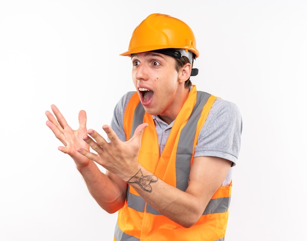 Hombre joven constructor de lado mirando sorprendido en uniforme aislado en la pared blanca