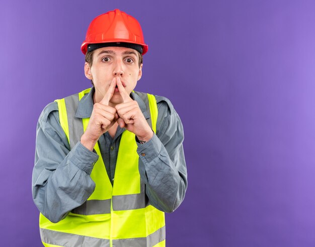 Hombre joven constructor sospechoso en uniforme mostrando gesto de silencio aislado en la pared azul con espacio de copia