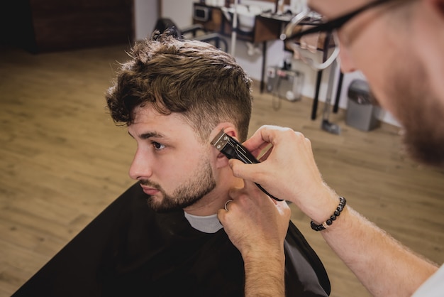 Foto hombre joven con corte de pelo de moda en peluquería. barber hace el corte de pelo y la barba.