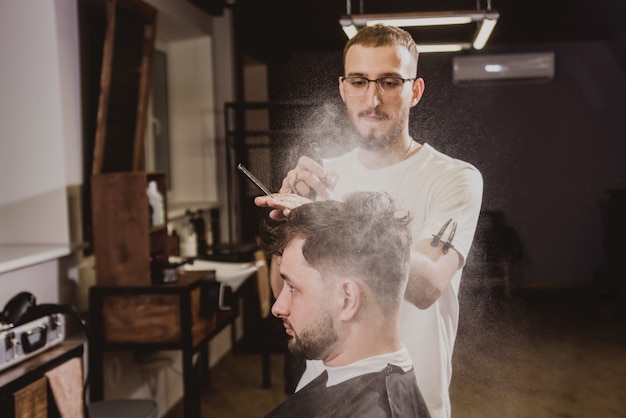 Hombre joven con corte de pelo de moda en peluquería. Barber hace el peinado y la barba.