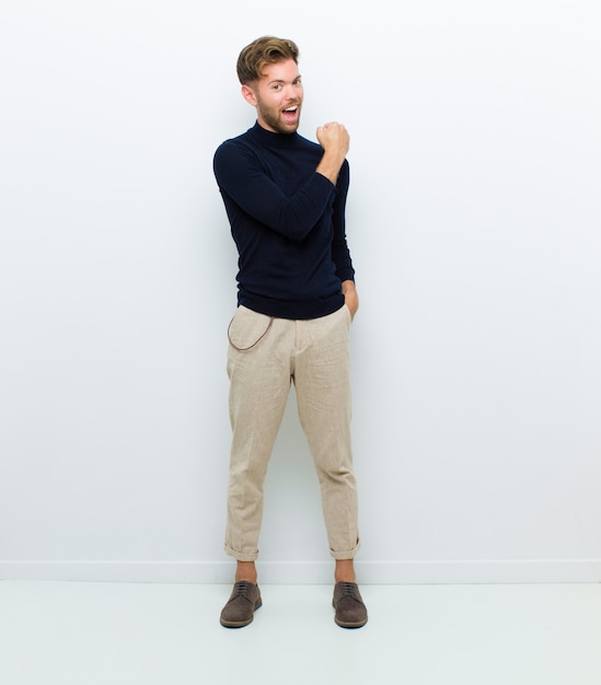 Hombre joven de cuerpo completo que se siente feliz, positivo y exitoso, motivado cuando enfrenta un desafío o celebra buenos resultados contra la pared blanca