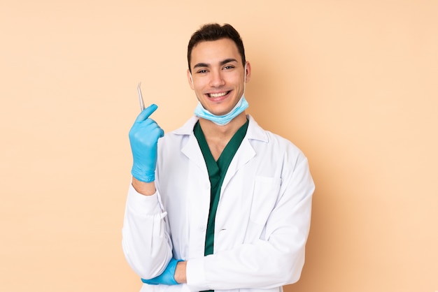 Hombre joven dentista con herramientas en color beige manteniendo los brazos cruzados en posición frontal