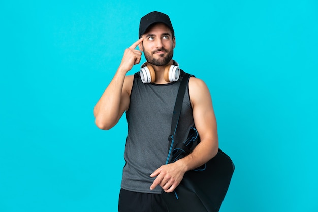 Hombre joven deporte con bolsa de deporte aislado en la pared azul con dudas y pensamiento