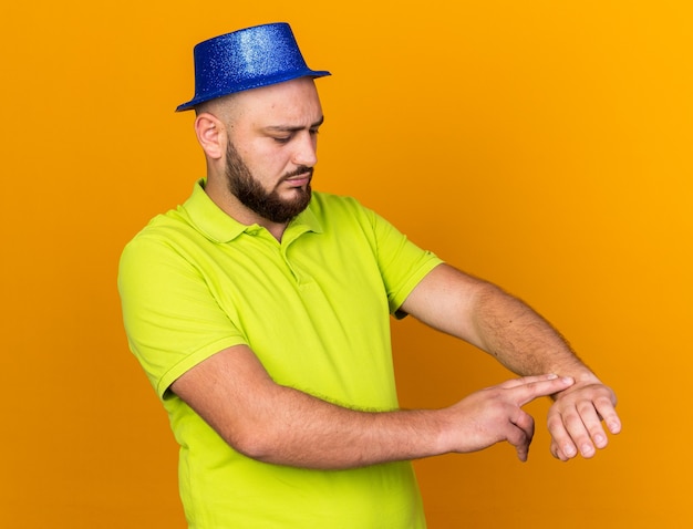 Hombre joven descontento con gorro de fiesta mostrando gesto de reloj de pulsera