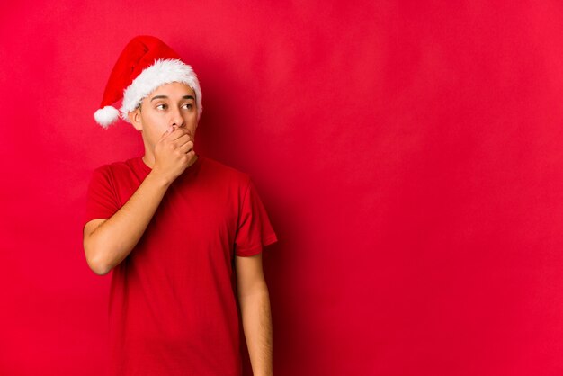 Hombre joven el día de Navidad pensativo mirando a un espacio de copia que cubre la boca con la mano.