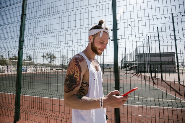 Un hombre joven con una diadema blanca en la cabeza y tatuajes en los brazos, vestido con la camiseta blanca, se encuentra junto a la valla del patio de recreo y usa el teléfono.