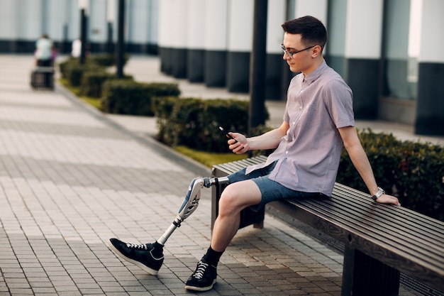 Hombre joven discapacitado con prótesis de pie sentado y sostener el teléfono móvil al aire libre