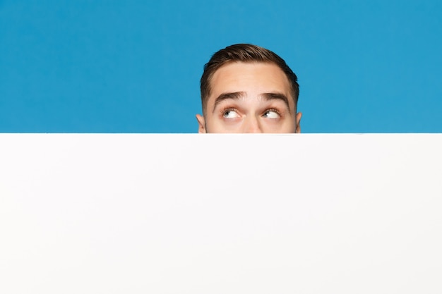 Hombre joven divertido que se esconde, mira por los ojos la gran cartelera en blanco vacía blanca para contenido promocional aislado en el retrato de estudio de fondo de pared azul. Concepto de estilo de vida de emociones de personas. Simulacros de espacio de copia.