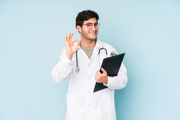 Hombre joven del doctor aislado en azul alegre y confiado que muestra gesto aceptable.