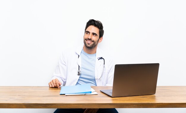 Hombre joven del doctor con su computadora portátil sobre la pared aislada que ríe y que mira para arriba