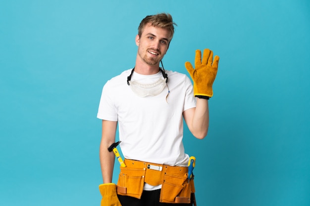 Hombre joven electricista sobre pared aislada saludando con la mano con expresión feliz