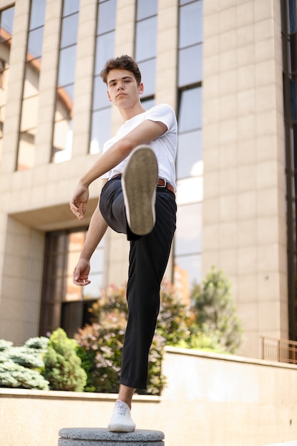 Hombre joven elegante con una camisa negra de moda en una camiseta blanca elegante con pantalones negros con un peinado de moda descansa cerca de un moderno centro de negocios. Chico atractivo en la calle en un día de verano.