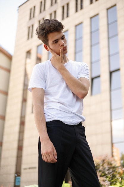 Hombre joven elegante con una camisa negra de moda en una camiseta blanca elegante con pantalones negros con un peinado de moda descansa cerca de un moderno centro de negocios. Chico atractivo en la calle en un día de verano.