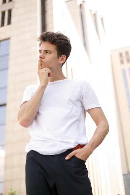 Hombre joven elegante con una camisa negra de moda en una camiseta blanca elegante con pantalones negros con un peinado de moda descansa cerca de un moderno centro de negocios. Chico atractivo en la calle en un día de verano.