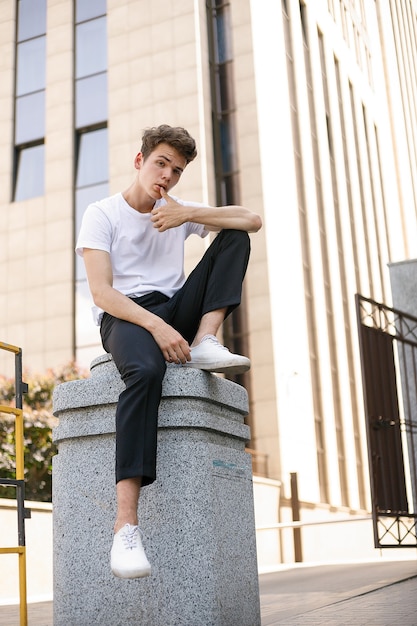 Hombre joven elegante con una camisa negra de moda en una camiseta blanca elegante con pantalones negros con un peinado de moda descansa cerca de un moderno centro de negocios. Chico atractivo en la calle en un día de verano.