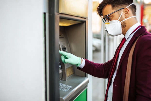 Hombre joven elegante con máscara protectora de pie en las calles de la ciudad y con máquina de cajero automático con guantes protectores en las manos. Concepto de salud y prevención de la pandemia de virus.