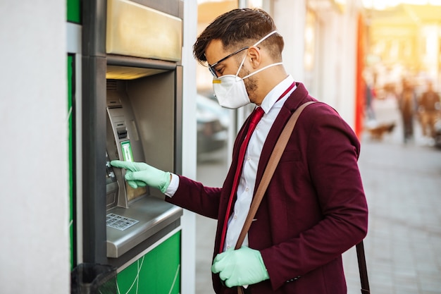 Hombre joven elegante con máscara protectora de pie en las calles de la ciudad y con máquina de cajero automático con guantes protectores en las manos. Concepto de salud y prevención de la pandemia de virus.