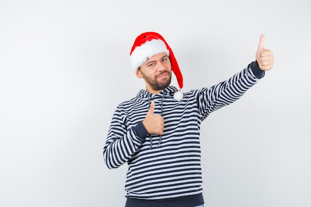 Hombre joven elegante que muestra los pulgares dobles para arriba en la sudadera con capucha, gorro de Papá Noel y que parece complacido.