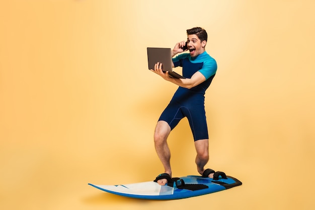 Hombre joven emocionado en tabla de surf hablando por teléfono usando la computadora portátil.