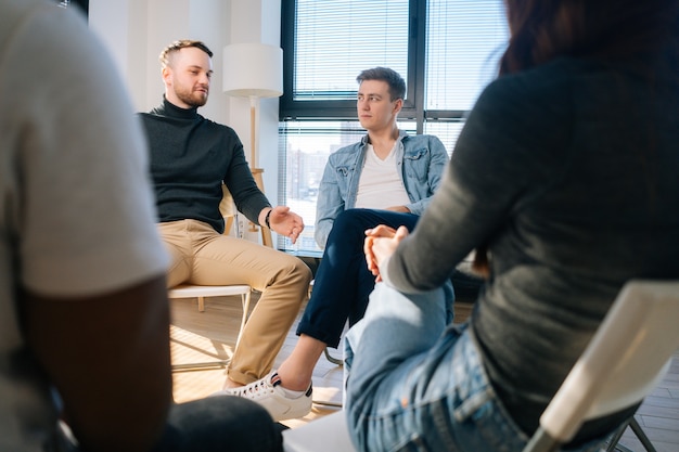 Hombre joven emocional que cuenta su problema mental o adicción a otros pacientes sentados en círculo. Concepto de consulta grupal de problemas de salud mental.